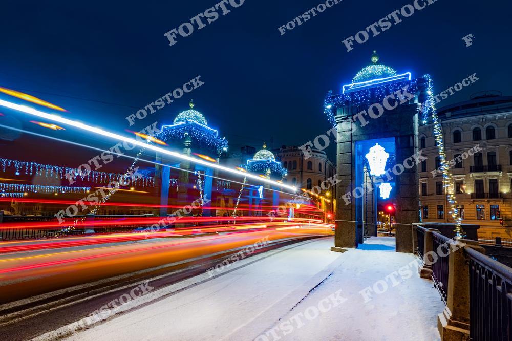 На фотографии изображен мост Ломоносова, украшенный яркими гирляндами и новогодними огнями. Архитектура моста с его каменными башнями выделяется на фоне динамичных световых следов транспорта. Зимняя ночь и праздничная подсветка создают неповторимую атмосферу. Этот мост – один из знаковых символов исторического центра Санкт-Петербурга.